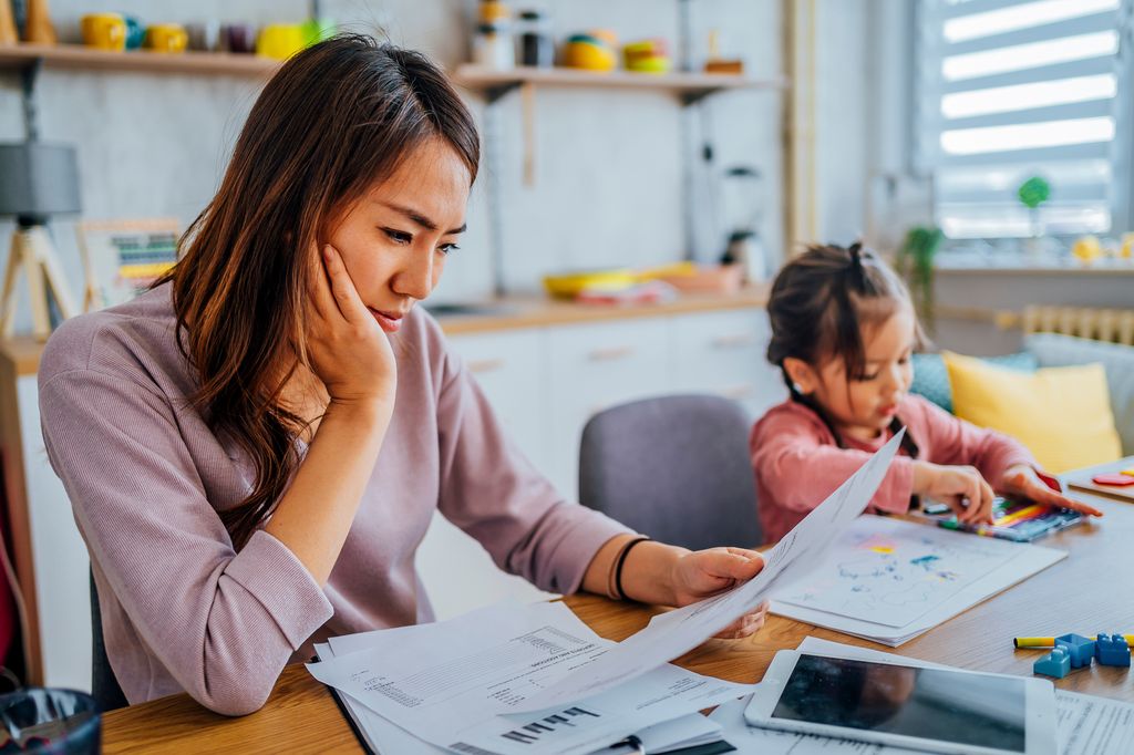 Young stressed mother checking her finances while her daughter is playing next to her