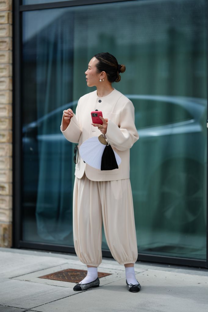 A guest wears dark hair slicked back into a low bun, pearl drop earrings, a collarless ivory jacket in a structured, soft drapey fabric with long wide sleeves, a chest patch pocket, a straight hem, and a concealed front finished with a single oversized dark button at the neckline, paired with matching ivory balloon trousers cut wide through the leg with deep pleats and elasticized cuffs, a small black drawstring pouch and a white pleated hand fan on a gold-tone ring handle are carried with a bright pink smartphone, white crew socks, and black ballet flats with a glossy cap toe and bow detail, standing still, outside Birrot, during Copenhagen Fashion Week