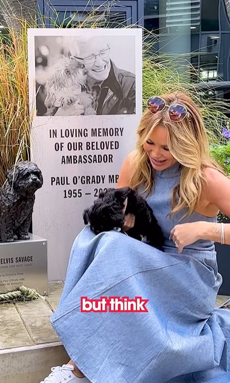 Amanda Holden with a black puppy in front of a memorial plaque for Paul O'Grady