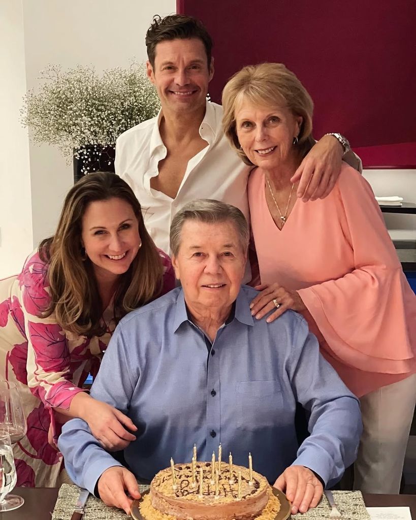 Ryan Seacrest stands next to his mom (R) and sister (L) as they stand behind his father Gary (seated) who has a birthday cake in front of him