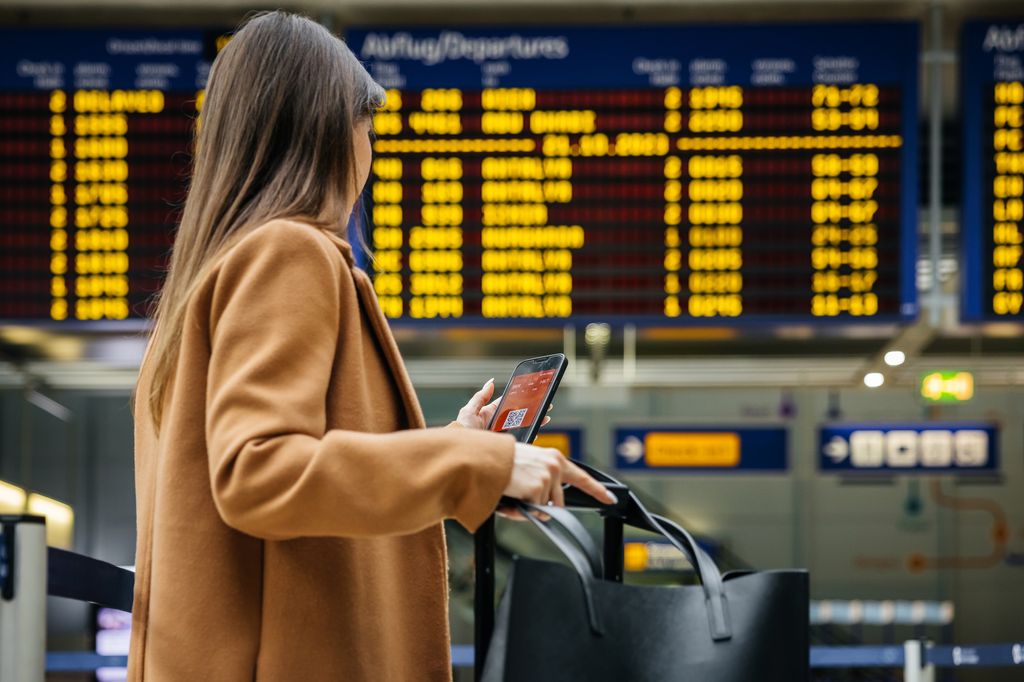 A woman traveler checks her mobile phone in front of the airport's flight information board, preparing for her upcoming solo journey.