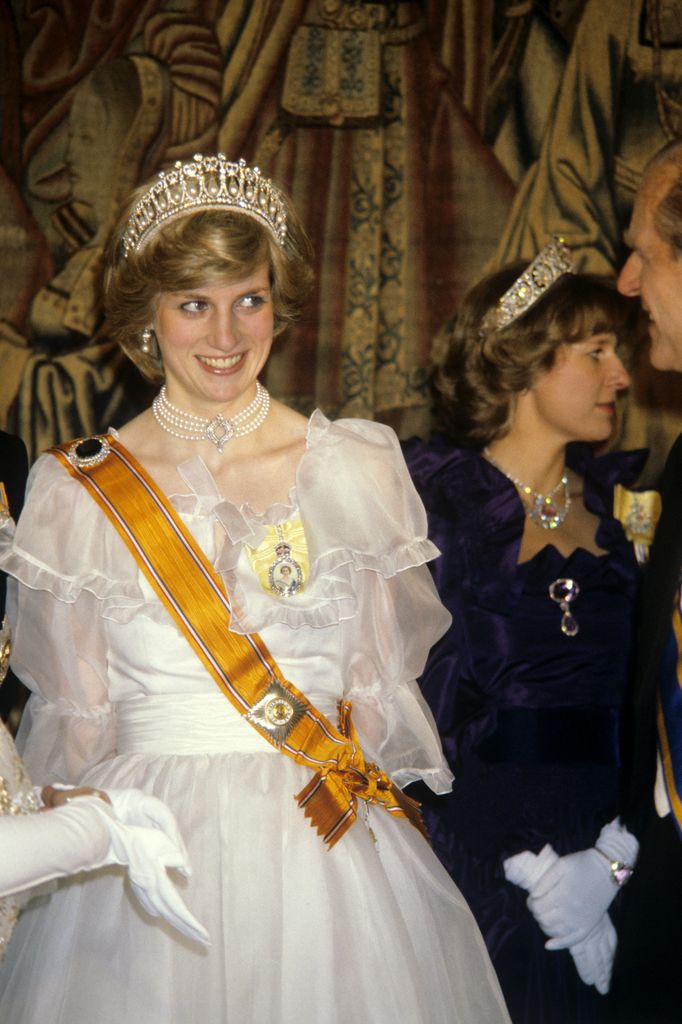 Princess Diana wears a white gown with a double-strand pearl choker, diamond tiara, and a royal sash. She smiles, capturing her elegance and charm at a banquet for the British royal family given by Queen Beatrix and Prince Claus of the Netherlands at Hampton Court Palace