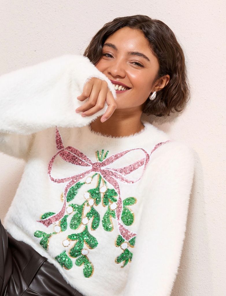 Next Christmas jumper for women with mistletoe on the front. The jumper is cream and it has a pink bow. The model is wearing with black leather trousers and statement earrings. 