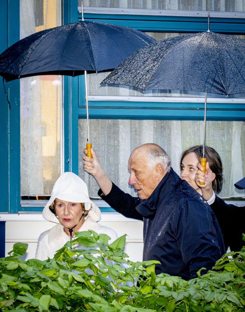 Queen Sonja in a white raincoat and King Harald in a blue raincoat underneath umbrellas