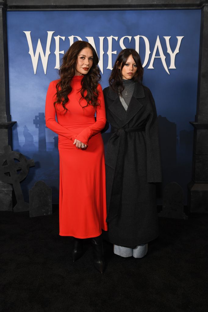 photo of catherine zeta jones in red dress next to jenna ortega in black trench coat