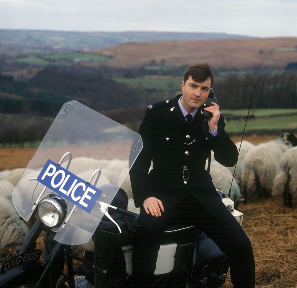 Nick Berry in a police uniform sitting on a motorbike with sheep around him