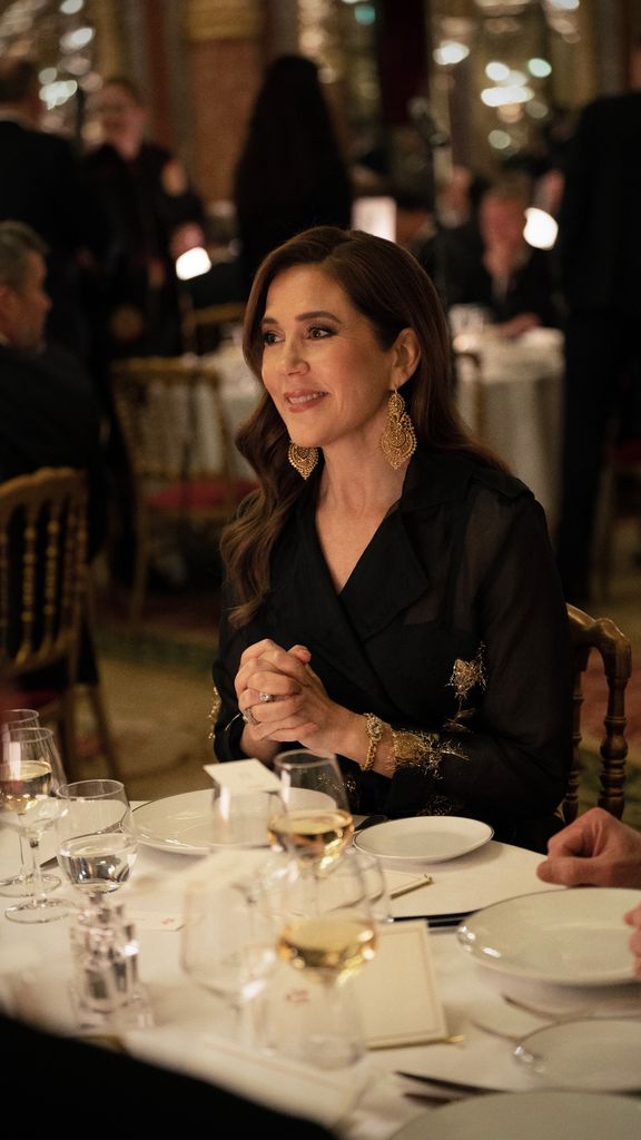 Queen Mary wearing black dress and gold jewellery, sitting at dining table