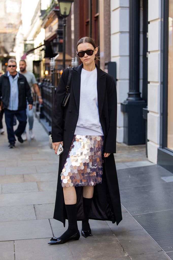  A guest wears sequined skirt, black long coat, Balenciaga bag, sunglasses, mid high socks outside Bora Aksu show