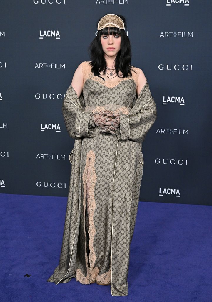  Billie stuns in a Gucci logo olive-green satin gown with lace trim. She accessorizes with a matching sleep mask on her head, soft black hair, and layered necklaces.