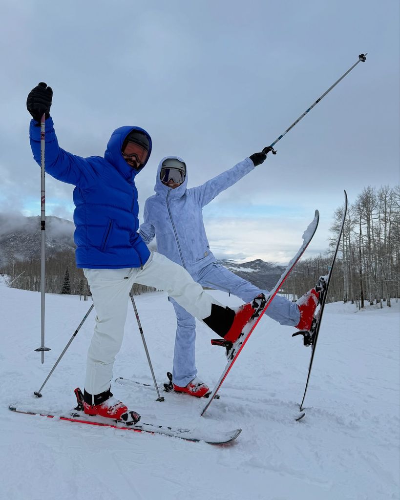 Heidi Klum and her partner in ski clothes on a mountain raising their legs in the air