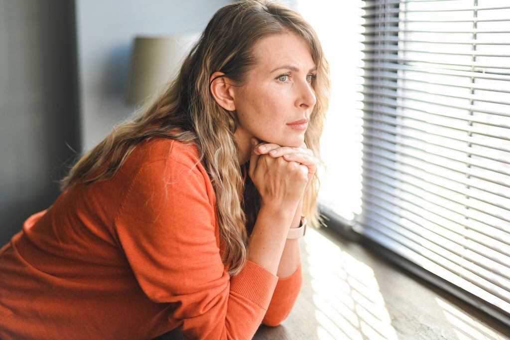 Mature adult woman in orange jumper looking out of the window wistfully