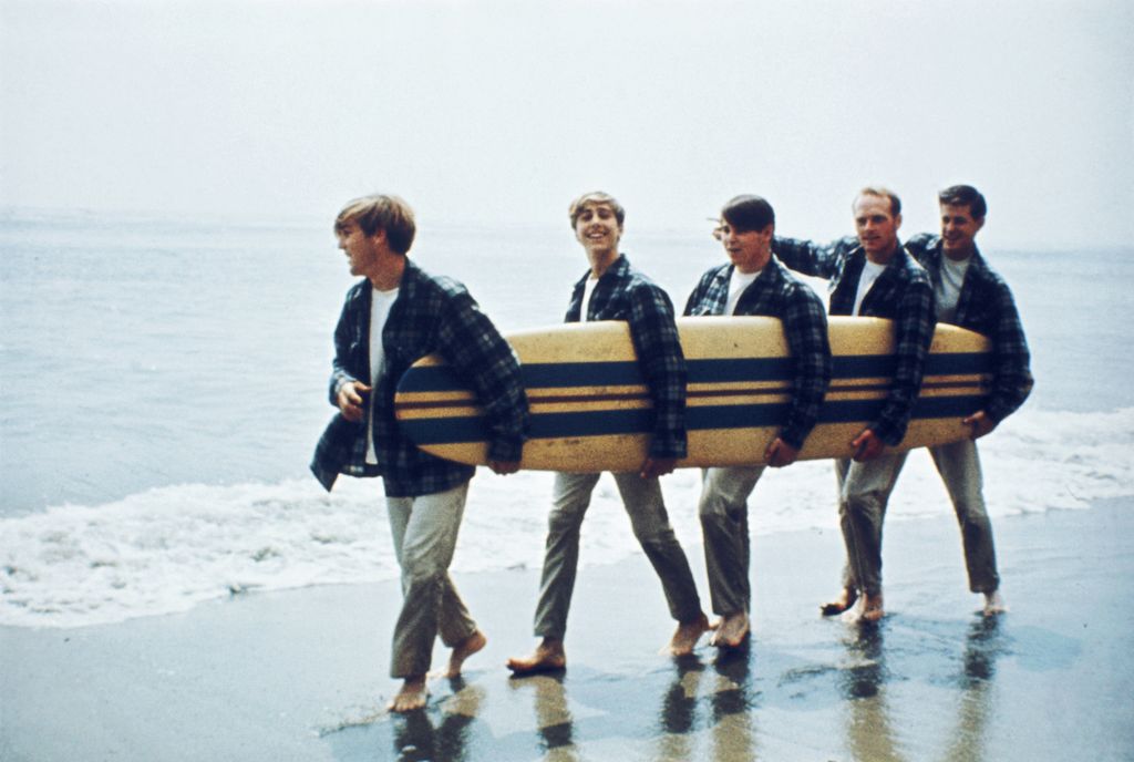 American Rock and roll band The Beach Boys walk along a beach holding a surfboard during a photoshoot for their debut album, 'Surfin' Safari', Los Angeles, California, August 1962. Left to right: Dennis Wilson, David Marks, Carl Wilson, Mike Love and Brian Wilson