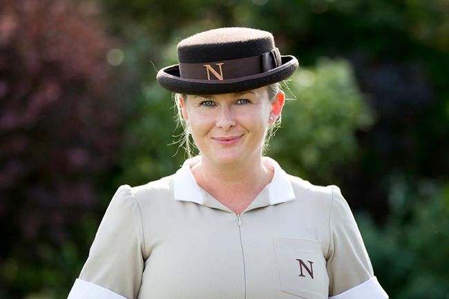 a photo of a woman smiling outdoors wearing a brown bowler hat marked with the letter n and a beige collared dress with the same n symbol on the chest