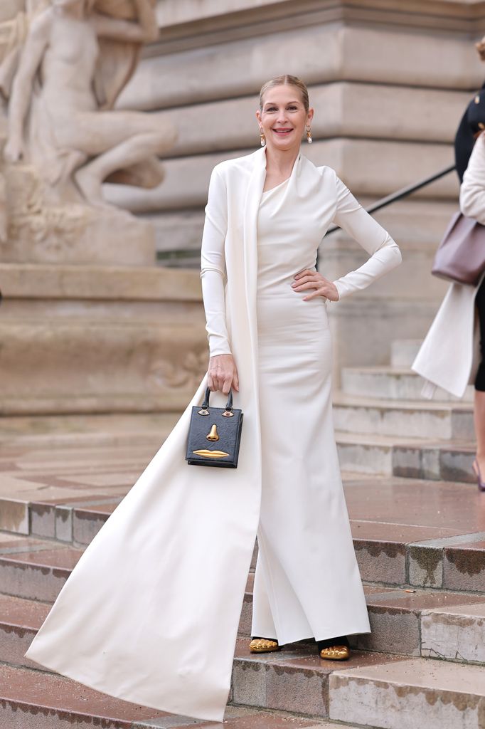 Kelly Rutherford wears a sleek white gown with a dramatic draped design was paired with a surrealist black handbag featuring a golden face motif. The look was accessorized with gold earrings and confidence.