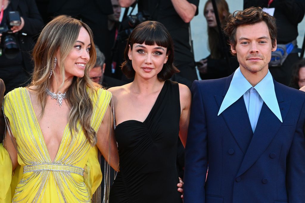 Olivia Wilde wears a vibrant yellow dress and glances over at Harry Styles, who is dressed in a blue suit. Between them, Sydney Chandler wears a black off-the-shoulder dress