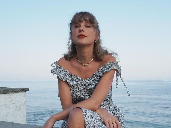 Taylor Swift sits on wall with sea behind her
