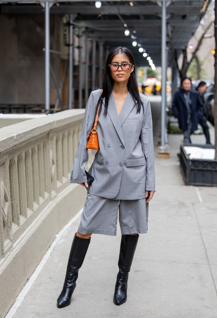 A guest wears grey wide shorts, oversized blazer, orange bag, black boots outside Dennis Basso during New York Fashion Week