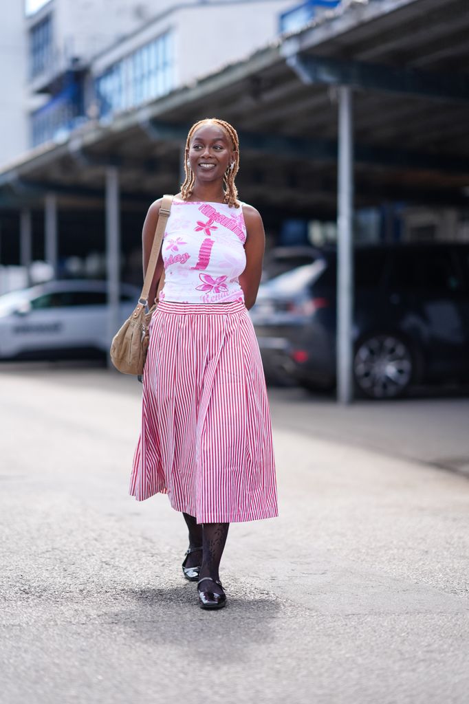 A guest wears silver earrings, white with bright pink pattern/print sleeve shirt, dark brown leather bag with strap, red with white lined pleated midi skirt, black tights, shiny black Ballerina flats leather shoes, outside Berner KÃ¼hl, during the Copenhagen Fashion Week 
