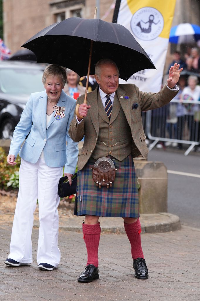 King Charles waves wearing kilt and holding umbrella