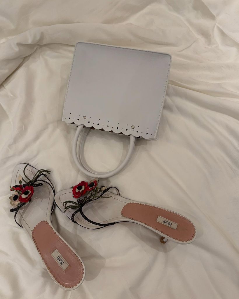 A pair of Prada kitten heels with red floral detail sit on the bed next to a white top handle handbag with scalloped detailing