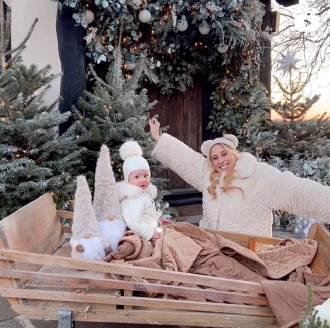 stacey kneels beside a wooden sleigh with her arms stretched out in joy as her toddler sits in the sleight beside two gnomes and they are wearing matching cream winter clothes