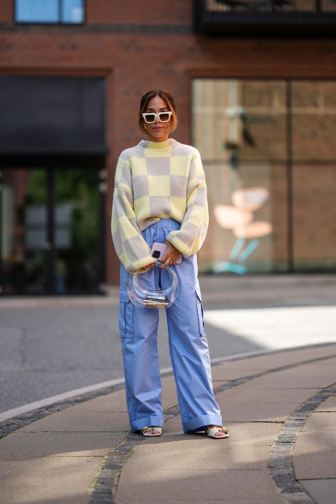 A guest wears white squared sunglasses, a pale yellow and pale gray checkered print pattern wool pullover, pale blue silk satin cargo wide legs pants, a white transparent plastic handbag, gold rings, white latte matte leather with a gold buckle heels mules , outside Lovechild 1979 , during the Copenhagen Fashion Week Spring/Summer 2024 on August 08, 2023 in Copenhagen, Denmark. (Photo by Edward Berthelot/Getty Images)