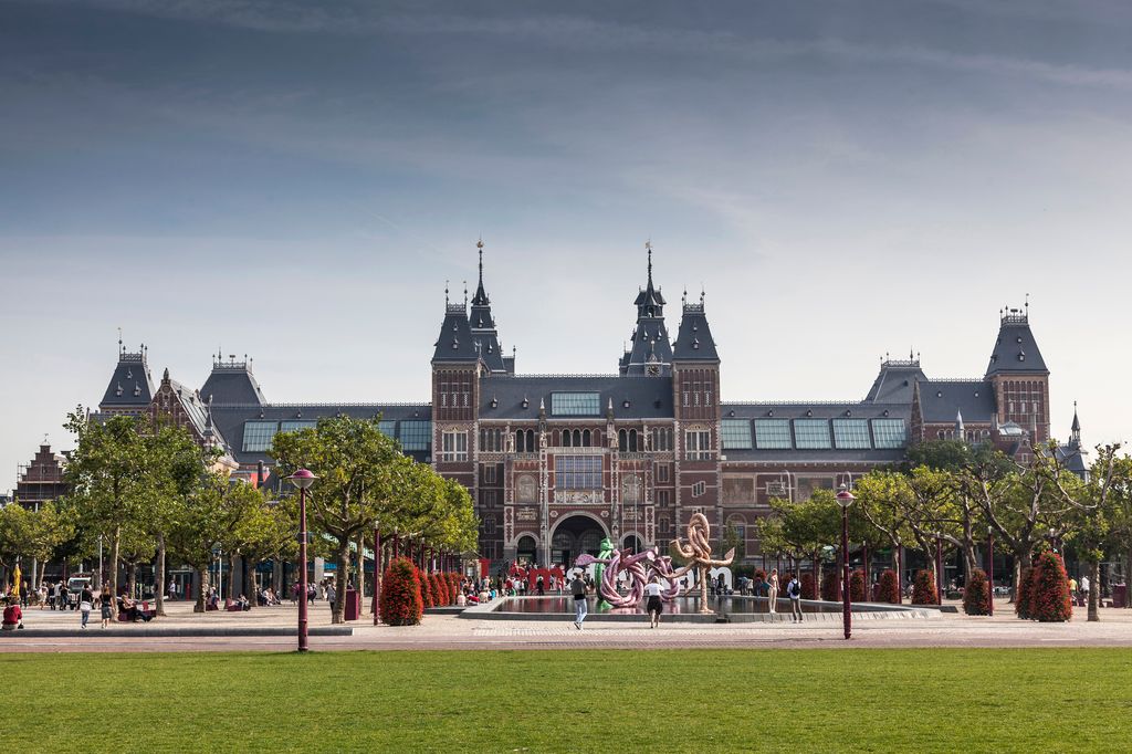Exterior view of Amsterdam’s Rijksmuseum, showcasing its historic Dutch architecture with a large central arch, ornate brickwork, and surrounding gardens under a cloudy sky