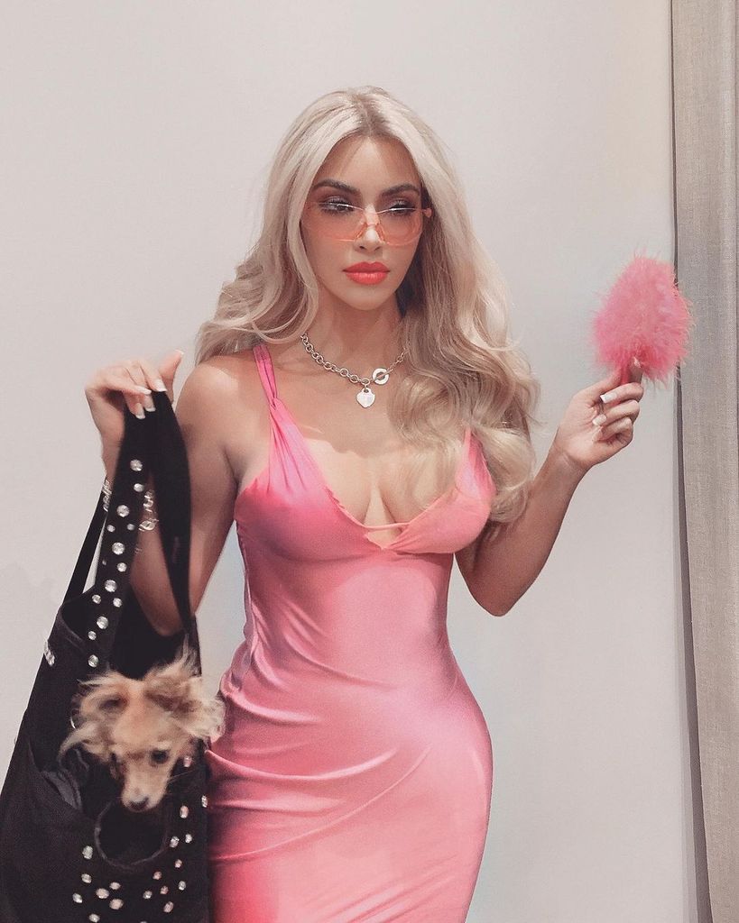 The image shows Kim Kardashian dressed as Elle Woods. She wears a tight, pink dress, blonde wig, pink sunglasses, and carries a black bag with a small dog inside, holding a fluffy pink pen.