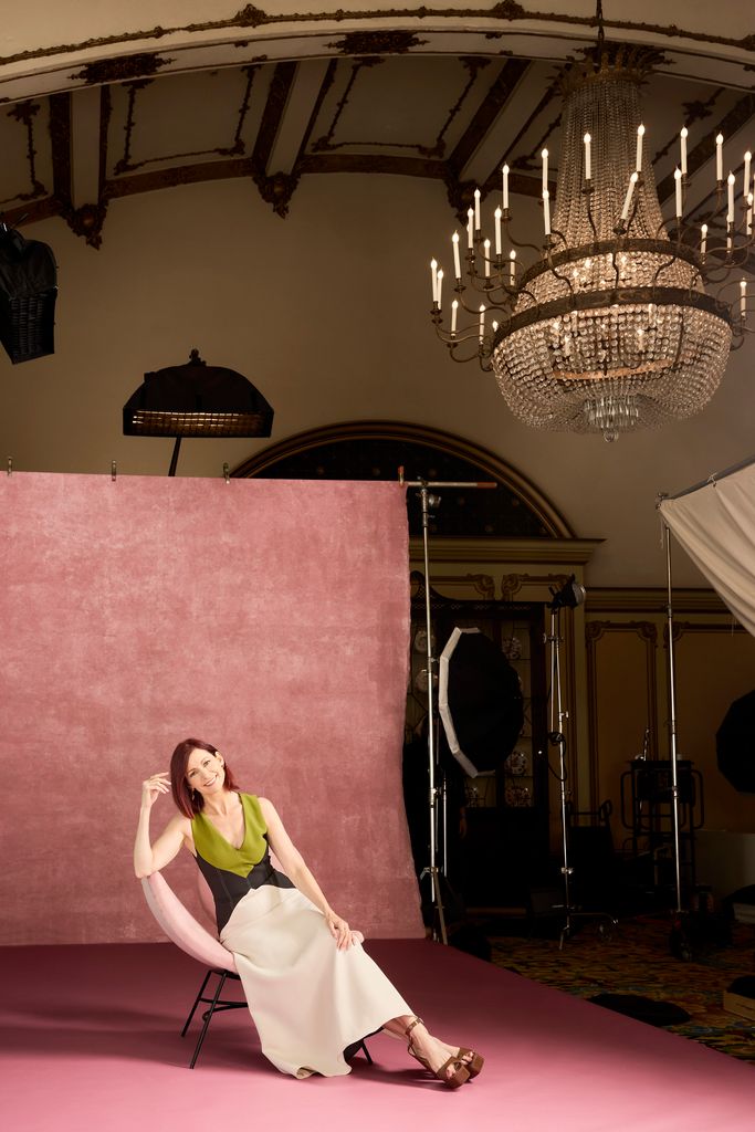 Carrie Preston sits on a pink chair with a large chandelier on the ceiling above her