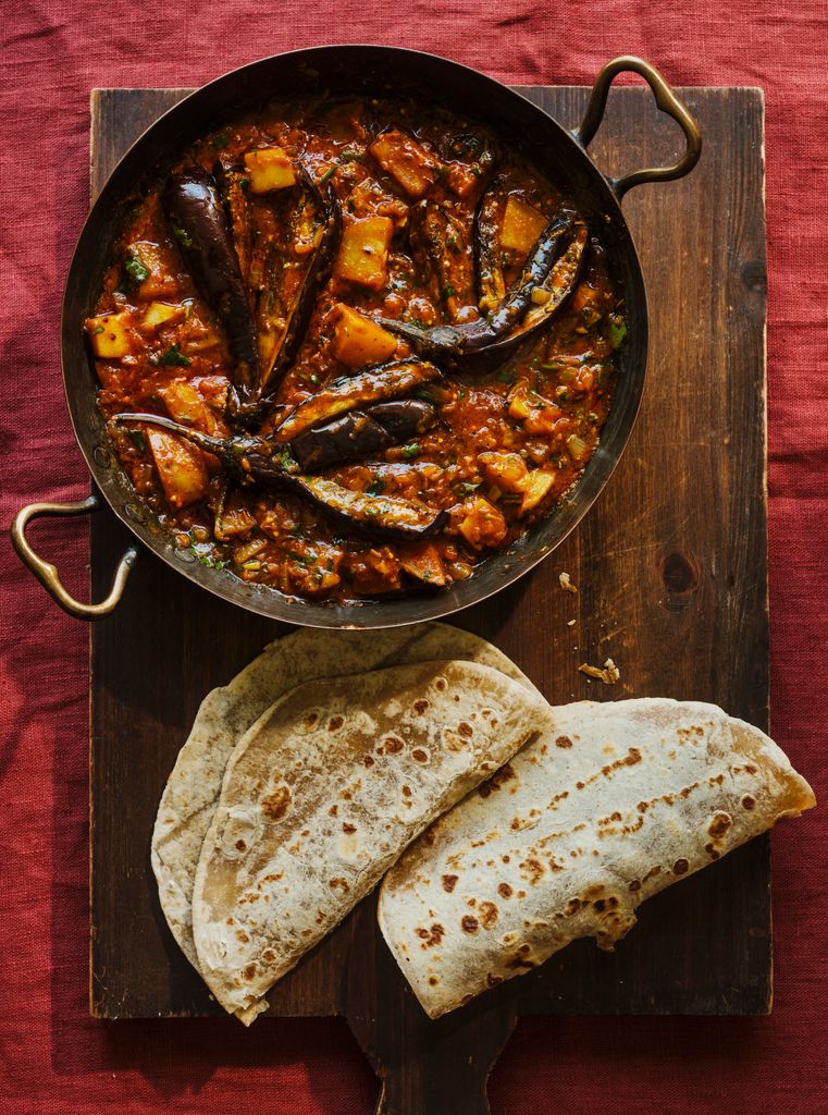 A wooden chopping board with an aubergine dish on top and flatbread on the side 
