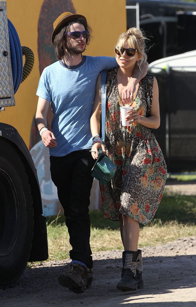 Sienna Miller and Tom Sturridge at Glastonbury 2013. Tom is wearing a blue t-shirt and tan hat while Sienna wears a printed floral dress and studded ankle boots. 