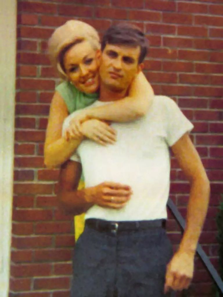 a younger dolly parton standing behind carl dean with her arms around his neck