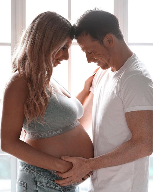 in profile stacey wears a cropped light grey top and joe who is wearing a white t shirt stand facing each other with their foreheads touching and they smile happily as they look down at staceys bare baby bump