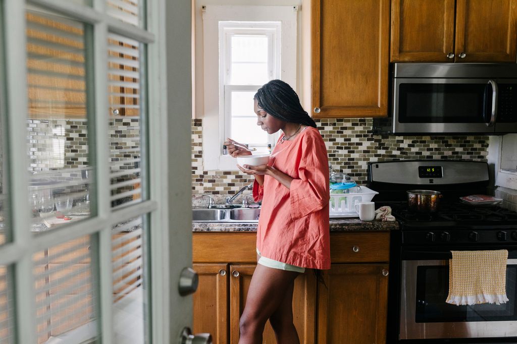 A woman wearing an oversized t-shirt and shorts standing in her kitchen and eating a bowl of cereal.