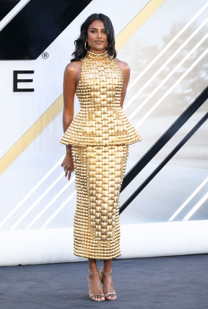 Simone Ashley, a British-Indian actress with long, softly waved black hair, stands poised and confident against a modern monochrome and gold backdrop. She wears a figure-hugging, futuristic gold dress that appears to be constructed from small, metallic, rectangular tiles—each catching the light like a jewel. The structured dress flares at the waist with a peplum detail, enhancing her hourglass silhouette, and extends into a form-fitting pencil skirt that ends mid-calf. Her neckline is high and halter-style, lending a regal air to the ensemble. She accessorizes with large gold hoop earrings and gold strappy heels that perfectly complement the ensemble’s opulence. Her makeup is minimal but luminous—bronzed skin, soft smoky eyes, and a neutral lip—allowing the dress to command center stage.