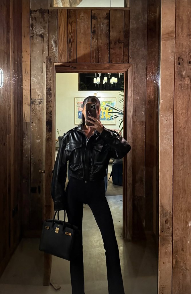 Maura Higgins takes a mirror selfie in a wooden panelled room wearing a black leather jacket, flared leggings and a black handbag.