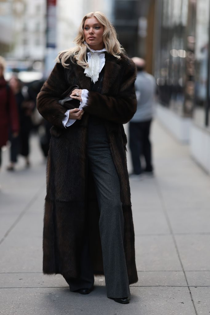 Elsa Hosk seen wearing dark brown full-length fur coat draped over a romantic white ruffled blouse, tailored charcoal grey waistcoat, dark grey wide-leg suit trousers, Ralph Lauren black leather clutch bag and black leather pointed boots, outside the Ralph Lauren Show, during New York Fashion Week, on September 10, 2025 in New York City