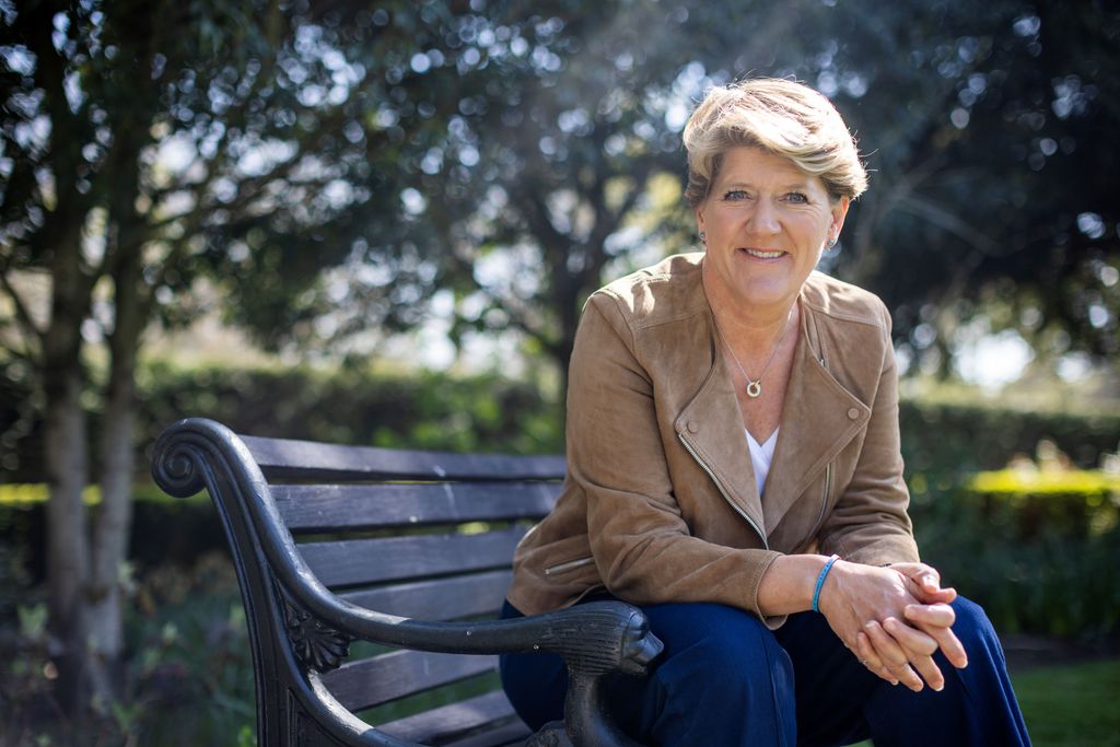 Clare Balding in suede jacket and jeans posing on park bench