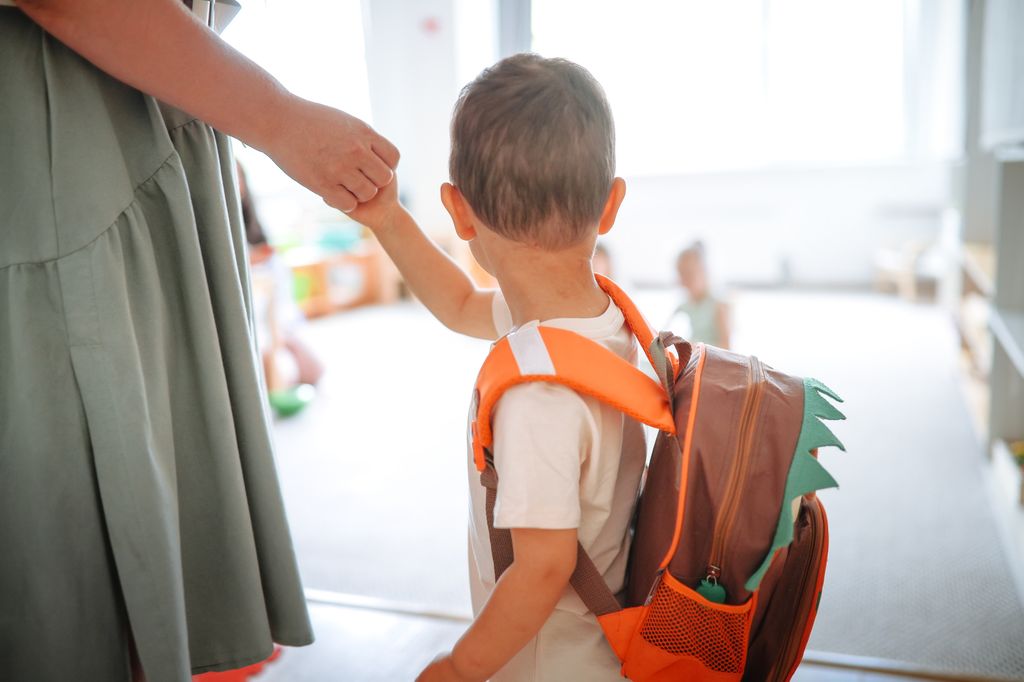Young child with a backpack holds mom's hand arriving at kindergarten, adapting to the children's group