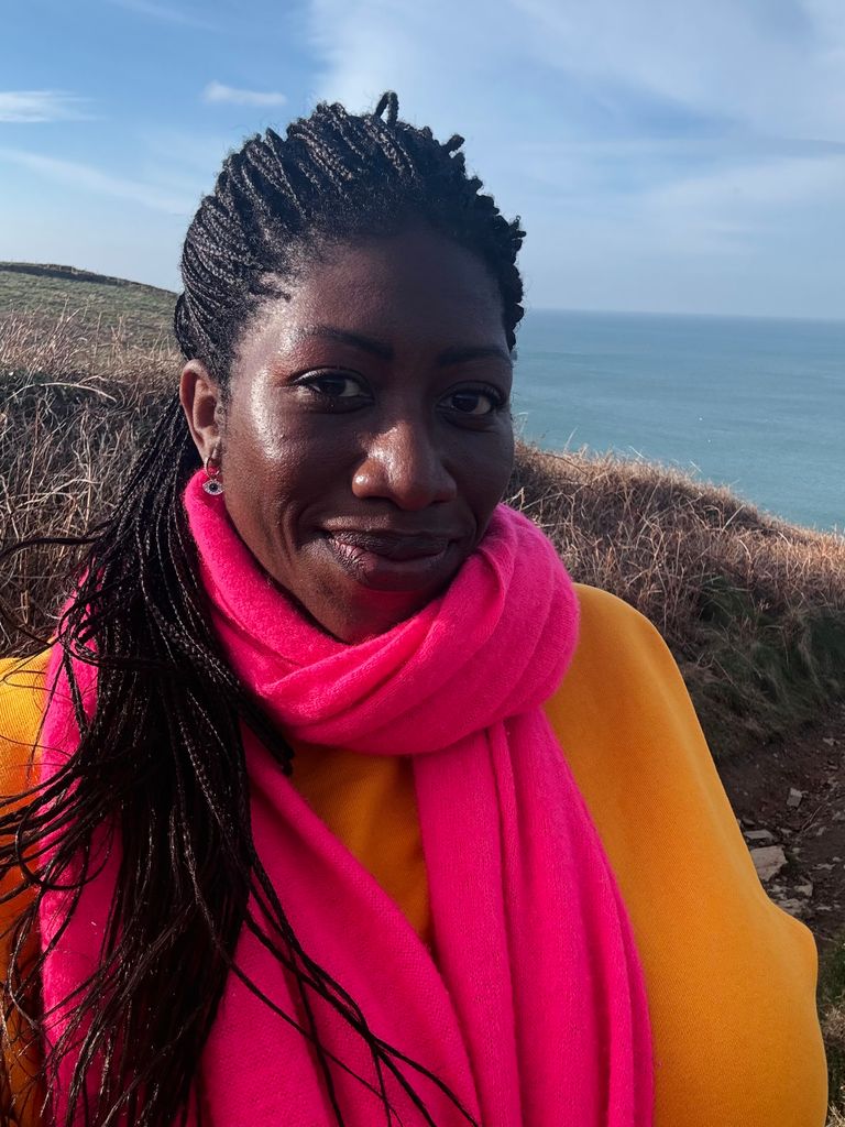 Ateh Jewel on the coast in a pink scarf and orange jumper smiling for the camera
