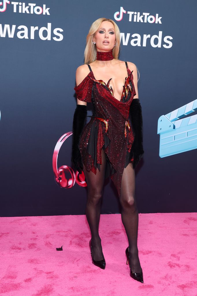Paris Hilton attends the TikTok Awards 2025 and poses on the pink carpet in a black and red sequin ensemble with matching choker, fishnet tights and black heels. 