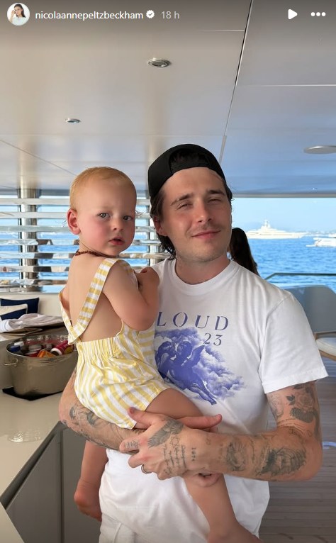 Brooklyn smiling for camera, holding up a toddler in a photo posted on Instagram. Brooklyn appears to be on a boat with the blue ocean in the background