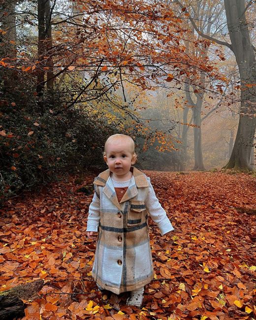 a toddler manages to stand up straight unaccompanied in a forest scattered with bright orange leaves while wearing a smart warm looking cream and brown check coat