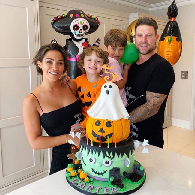 a man and woman each hold a child up high so that they can be seen over the top of a large cake which is decorated to look like a white ghost on top of an orange halloween pumpkin on top of a green frankenstein head