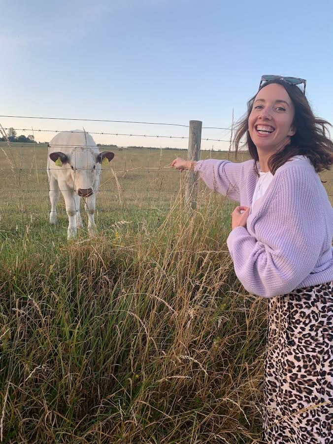 Young woman smiling widely in a purple cardigan as she reaches for a cow