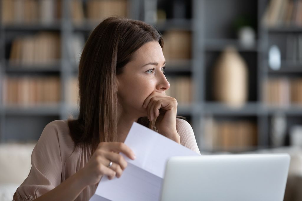 Head shot stressed young woman holding paper document, bank debt notification, thinking of financial troubles, looking away. Lost in negative thoughts depressed woman worrying about bad news notice.