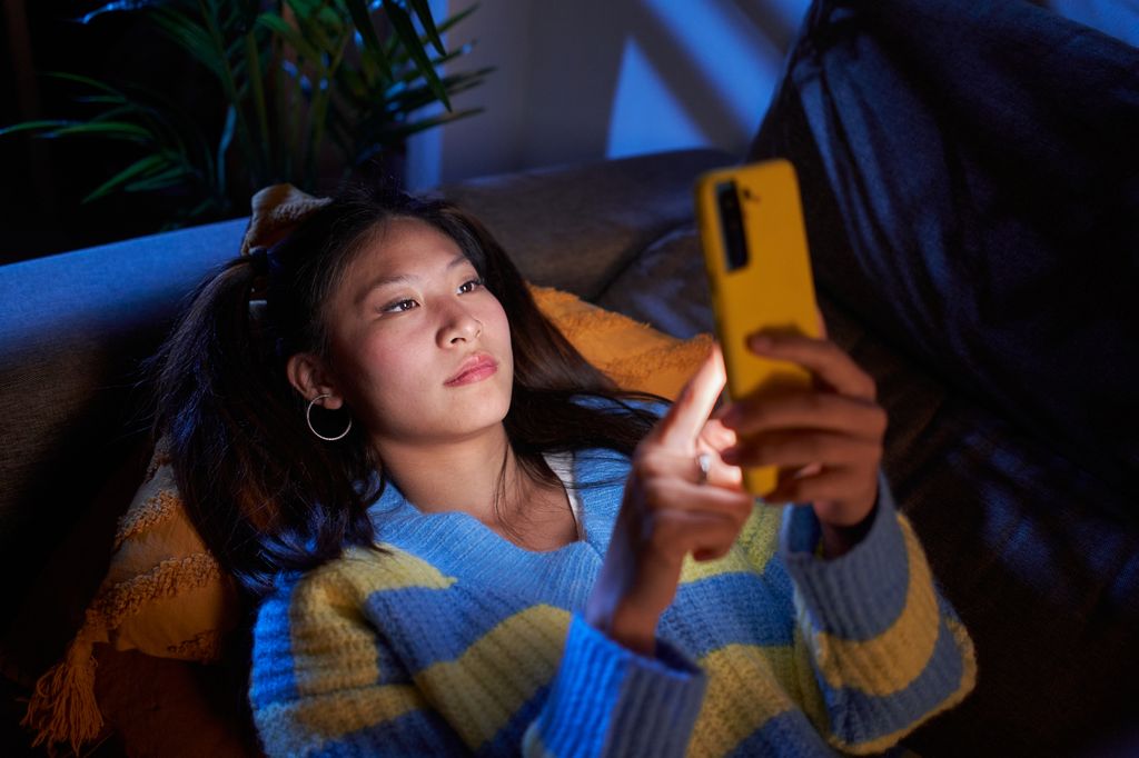 Young beautiful Chinese woman relaxing lying on the couch at home and texting on smartphone.