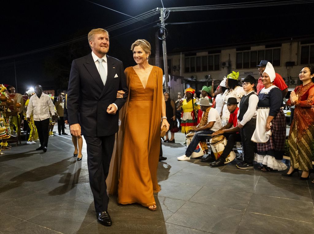 King Willem-Alexander and Queen Maxima in orange gown walking down dark street lined with people
