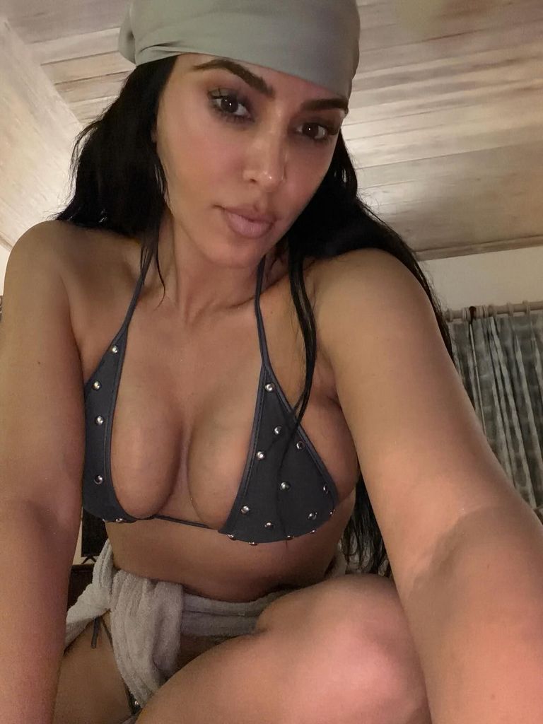 Kim Kardashian takes a selfie wearing a dark grey studded triangle bikini and a soft grey bandana on her head.