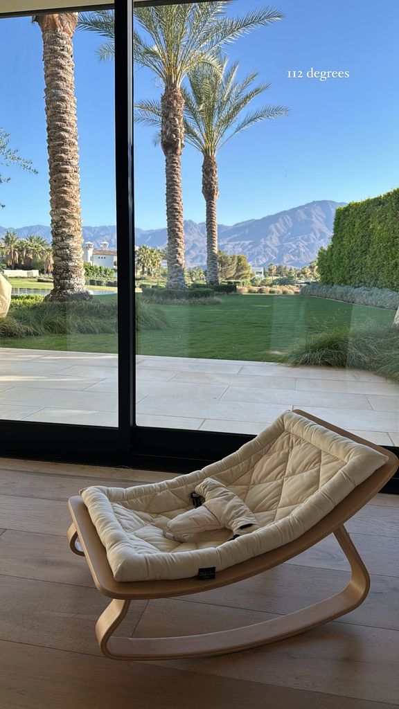 Picture posted by Kourtney Kardashian. A white rocker chair sits inside on wooden floor, and outside the floor to ceiling glass windows are palm trees